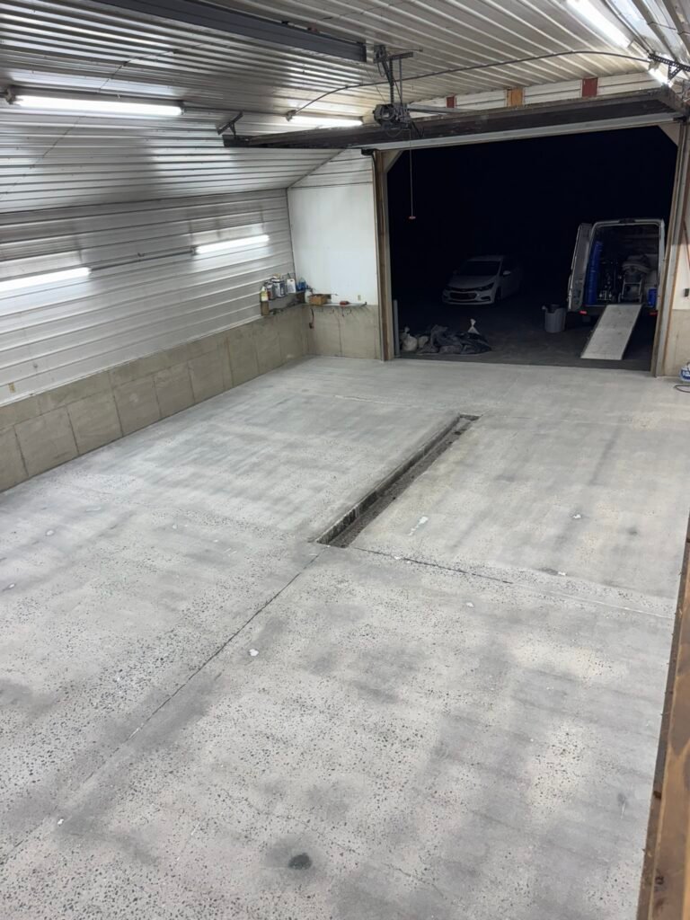 Bare concrete pole-barn floor before coating, garage door open at night with vehicles outside